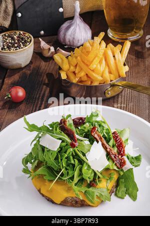 Cibo, delizioso cheeseburger condito con rucola, pomodori secchi su un tavolo rustico con contorni Foto Stock