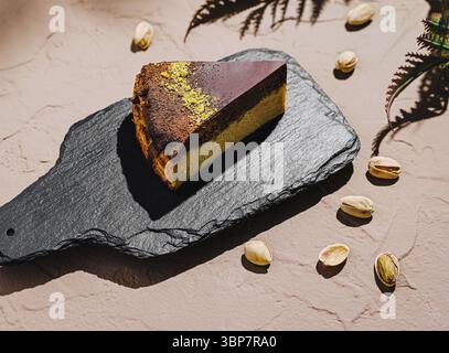 Cibo, Una fetta di torta a strati ben presentata si trova su un piatto di ardesia, circondato da pistacchi sparsi. Il ricco dessert presenta un cioccolato Foto Stock