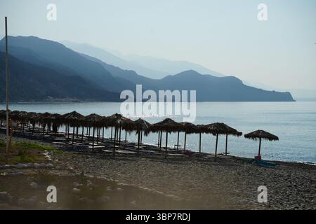 Tranquilla scena costiera con file di ombrelloni di paglia su una spiaggia di ciottoli, che si affaccia sulle acque calme e sulle montagne lontane durante il tramonto, creando un ambiente tranquillo Foto Stock
