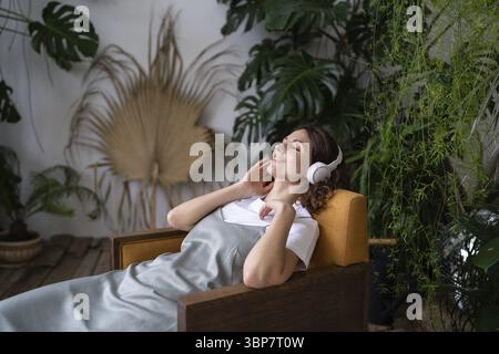 Bella giovane e sognante donna con gli occhi chiusi seduta su una poltrona con cuffie wireless e ascolta musica o podcast tra ev esotici Foto Stock