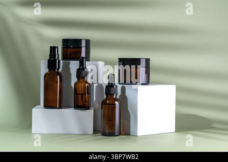 Set di prodotti di bellezza in bottiglie e vasi in vetro ambrato su podi bianchi su sfondo verde con ombra di foglie di palma. Set per la cura della pelle per la routine quotidiana. Mo Foto Stock