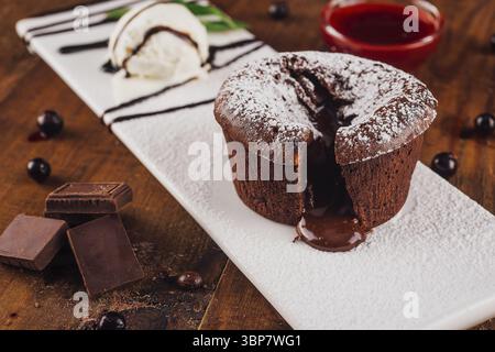Cibo, torta lavica al cioccolato che trasuda cioccolato fuso su un piatto bianco, servito con una paletta di gelato alla vaniglia, frutti di bosco e pezzi di cioccolato Foto Stock