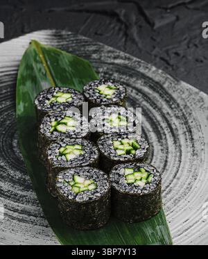 Cibo, sushi involtini di riso nero ripieni di cetriolo fresco sono disposti ordinatamente su un piatto a motivi geometrici. La presentazione artistica mette in risalto il vibrante gre Foto Stock
