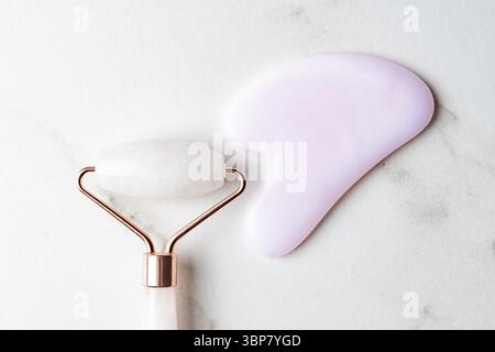 Rullo derma con massaggio in giada e pietra gua Sha su sfondo in marmo bianco. strumenti antinvecchiamento per la cura della bellezza per la routine quotidiana a casa Foto Stock