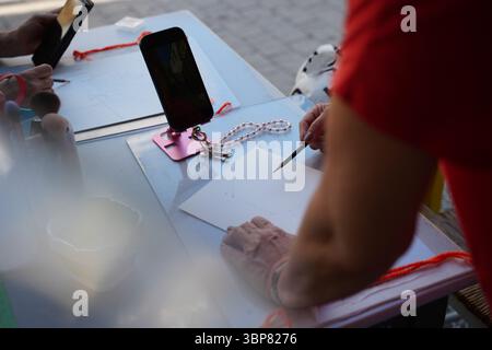 Un artista seduto a un tavolo all'aperto disegna su carta usando una matita, con uno smartphone su un piedistallo come riferimento. La scena cattura creatività e Foto Stock