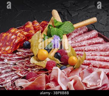 Cibo, Un piatto ben organizzato presenta una varietà di salumi, tra cui salame e prosciutto, accompagnati da frutta fresca ed erbe aromatiche. Perf Foto Stock