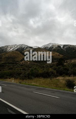 Maestose montagne innevate si stagliano sotto un cielo nuvoloso, una strada tortuosa attraversa una vegetazione lussureggiante, evocando avventura, serenità e natura Foto Stock