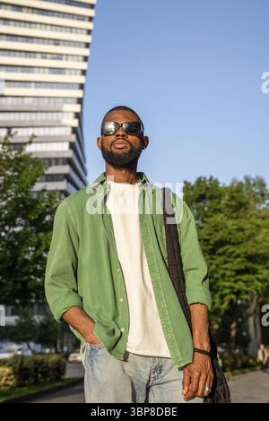 Elegante e serio uomo afro-americano che indossa occhiali da sole look sicuro in piedi con la mano in tasca sui jeans su un moderno sfondo grattacielo. Auto-assur Foto Stock