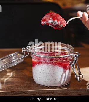 Cibo, tenere a mano un cucchiaio con budino di chia e marmellata di fragole su un barattolo su un tavolo di legno Foto Stock
