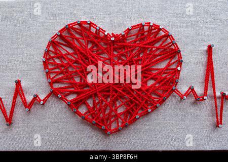 Archi artistici artigianali a forma di cuore con linee cardio a impulsi in filo rosso. Cuore di lana rossa, simbolo dell'amore, realizzato in filati di lana rossa aggrovigliati su chiodi metallici su sfondo tela Foto Stock