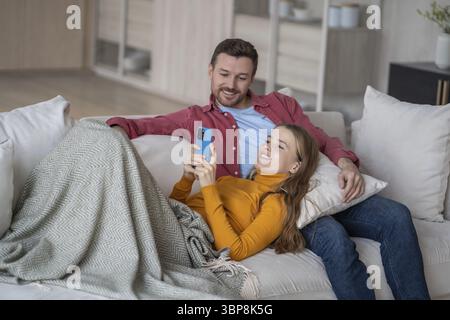 Uomo donna felice che si rilassa sul divano. Moglie sdraiata in grembo al marito usando lo smartphone, sorridente. L'uomo che ride guarda lo schermo del telefono. I coniugi guardano i rulli, sh Foto Stock