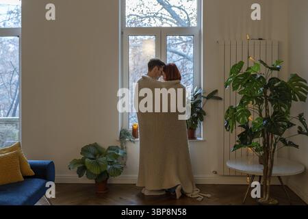 Coppia vista posteriore avvolta in una coperta che guarda dal finestrino di casa. Riscaldamento familiare a plaid. Moglie e marito si godono un momento tenero, sognando il futuro Foto Stock