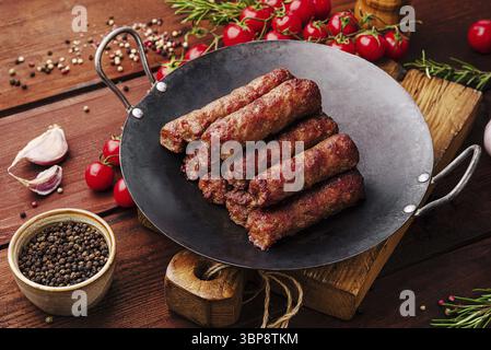 Cibo, succose salsicce di carne alla griglia sono impilate su una padella nera. Pomodori freschi, aglio ed erbe circondano il piatto, creando un'atmosfera rustica Foto Stock