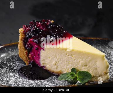 Cibo, fetta di cheesecake cremosa condita con una vivace composta di bacche e zucchero a velo, servita elegantemente su un piatto scuro Foto Stock