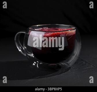 Cibo, Una moderna tazza di vetro a doppia parete piena di tè rosso, isolata su uno sfondo scuro e umido Foto Stock