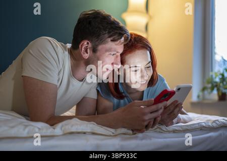 Coppia che guarda il telefono mentre si sdraia a letto. Ragazza che mostra video divertenti del fidanzato al telefono. Marito e moglie felici che controllano i social media. Donna che mostra un uomo Foto Stock