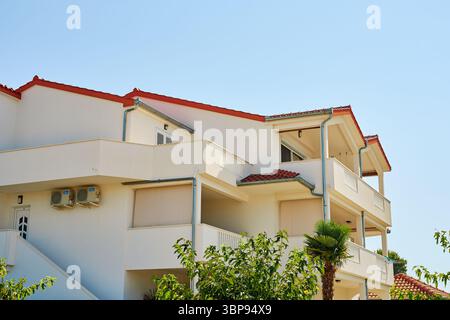 Edificio residenziale bianco con balconi, tetto piastrellato rosso e finestre aperte sotto il cielo blu. Facciata di una villa moderna con terrazze aperte e palme. Io Foto Stock