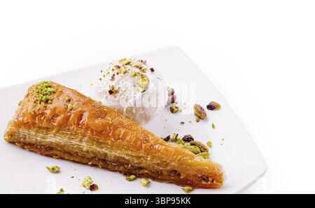Cibo, Golden baklava si trova elegantemente su un piatto bianco accanto a una paletta di gelato alla vaniglia, decorato con pistacchi schiacciati. Questo delizioso dessert Foto Stock