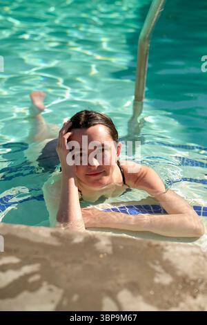 Donna che si rilassa in piscina, appoggiata su un gradino, con acqua cristallina intorno a lei. Palm Springs, Stati Uniti Foto Stock