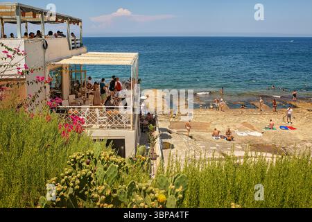 Sliema, Malta - 06/07/2025: Una vista attraverso una lussureggiante vegetazione fino ad un affollato caffè a due piani e alla spiaggia della città con piscine di roccia naturale e turisti Foto Stock