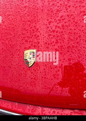 Primo piano di un cofano o cofano auto Porsche 911 rosso bagnato con il logo Porsche ricoperto da gocce di pioggia che riflettono la luce Foto Stock