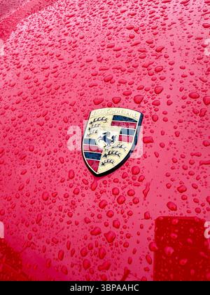 Primo piano di un cofano o cofano auto Porsche 911 rosso bagnato con il logo Porsche ricoperto da gocce di pioggia che riflettono la luce Foto Stock