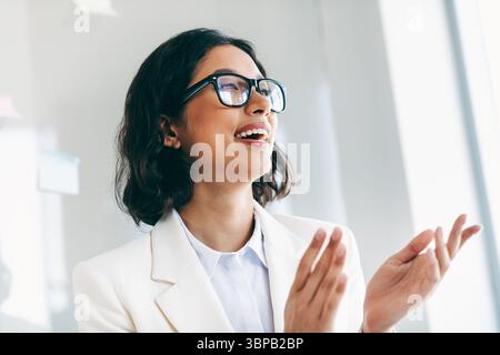 Affascinante donna d'affari in un moderno ambiente d'ufficio, che applaude ed esprime positività, vestita con un abito bianco professionale Foto Stock