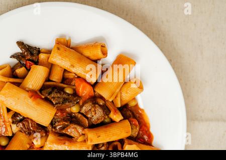 Stufato di manzo con pasta rigatoni, piselli, carote e funghi serviti su un piatto bianco, offrendo un pasto abbondante e saporito Foto Stock