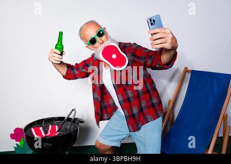 Uomo maturo che scatta selfie durante il barbecue con bistecche e birra in mano, godendosi l'estate all'aperto Foto Stock