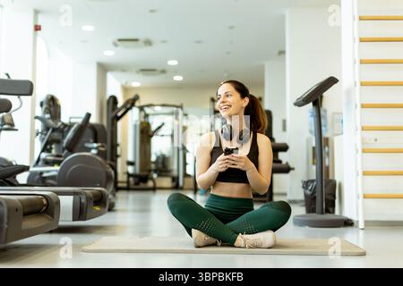 Sorridendo entusiasticamente tenendo le cuffie, una giovane donna gode di un momento di beatitudine in un vivace centro fitness. Foto Stock