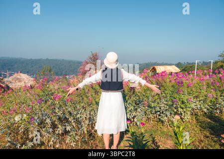 Il retro di una donna in abito bianco e cappello si erge a braccia aperte in un vivace campo di fiori rosa e si affaccia sulla vista panoramica delle montagne sotto il cielo azzurro cristallino del paese Foto Stock