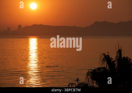 il vivace e suggestivo tramonto si dispiega sull'oceano calmo, con il grande sole che proietta un brillante riflesso dorato sull'acqua. silhouette distante Foto Stock