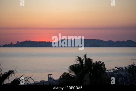la vibrante alba proietta calde sfumature sul tranquillo mare adriatico, con la costa sagomata di durazzo, albania, visibile in lontananza. palme fra Foto Stock