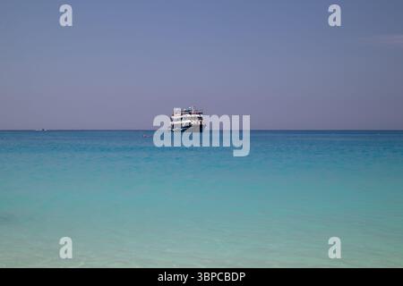 la piccola barca passeggeri galleggia tranquillamente all'orizzonte di un vasto mare turchese, con un cielo azzurro e quasi nuvoloso sopra, suggerendo un tranquillo e tranquillo Foto Stock