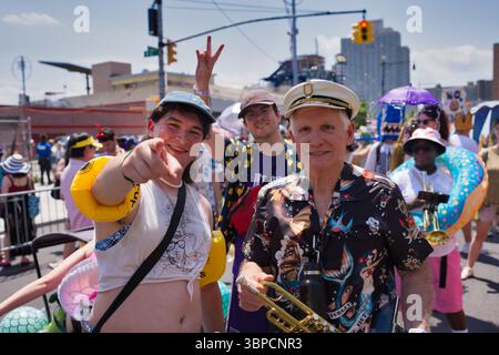 Una vivace scena di strada, forse una parata, presenta persone in abiti colorati. Un uomo con un cappello da capitano e una camicia floreale suona una tromba. Due youn Foto Stock