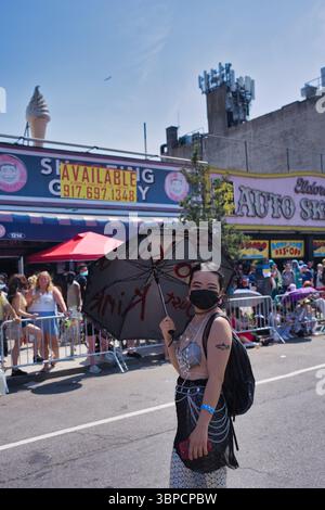Una giovane donna in stile sirena ha un ombrellone decorato. Indossa una maschera facciale e si trova in mezzo a una folla vicino ai negozi di Coney Island. La S Foto Stock