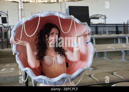 Una donna con i capelli castani ricci sorride mentre all'interno di un grande supporto rosa per conchiglie ornato da collane di perle. Indossa un corsetto beige e tiene i fili Foto Stock