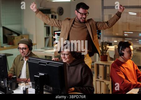 Gruppo di giovani che lavorano in postazioni di computer indossando cuffie nel call center truffa con un uomo caucasico che festeggia dietro di loro Foto Stock