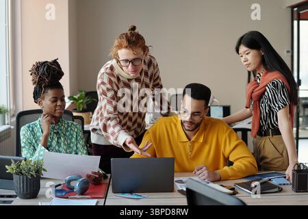 Donna nera che tiene i documenti seduti accanto all'uomo caucasico con gli occhiali che puntano al laptop mentre giovane adulto uomo nero e donna asiatica collaborano Foto Stock