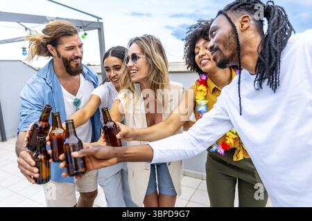 Amici felici che festeggiano sul tetto, brindano con bottiglie di birra e si godono momenti di qualità insieme in una vibrante atmosfera estiva Foto Stock