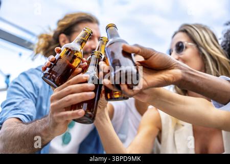 Gruppo di amici che festeggiano sul tetto, alzano le bottiglie di birra in alto per un brindisi gioioso, incarnano felicità e solidarietà Foto Stock
