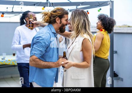 Coppia felice che balla gioiosamente e sorseggia birra in una vivace festa sul tetto, circondata da amici diversi che si godono una vivace serata estiva Foto Stock