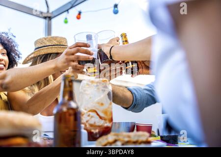 Gruppi diversi di amici che gusteranno cibo e bevande al barbecue, brindando insieme su un tetto sotto le luci a corde Foto Stock