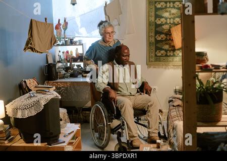 Donna caucasica anziana che assiste l'uomo Nero anziano seduto su sedia a rotelle in interni Foto Stock