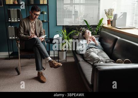 Giovane uomo che giace sul divano che parla con il terapista maschile Foto Stock