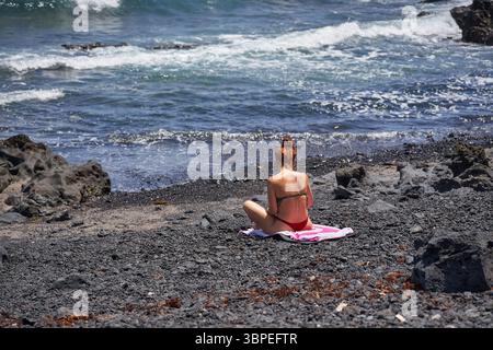Donna che prende il sole da sola su una spiaggia vulcanica vicino all'Oceano Atlantico a Malpaís de Güímar Foto Stock