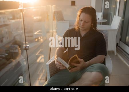 Uomo con i capelli bagnati che leggono un libro durante l'ora d'oro postura rilassata a piedi nudi seduto in rattan sedia balcone urbano. Foto Stock