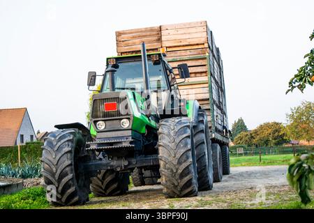 Grande trattore agricolo verde con rimorchio caricato con casse di legno, parcheggiato su un sentiero sterrato in un ambiente rurale con edifici agricoli e alberi. Foto Stock