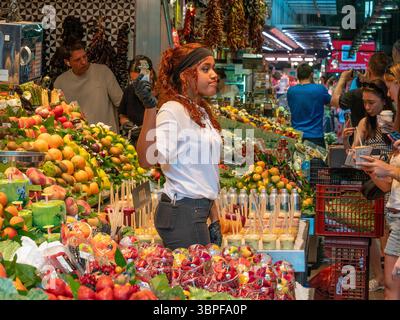 Un venditore serve succhi di frutta fresca e insalate ai clienti in una vivace e colorata bancarella di frutta all'interno del mercato la Boqueria, Barcellona, Spagna. Foto Stock