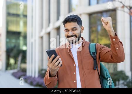 Un giovane eccitato guarda il suo telefono mentre festeggia le buone notizie all'aperto, alzando il pugno in trionfo. Foto Stock